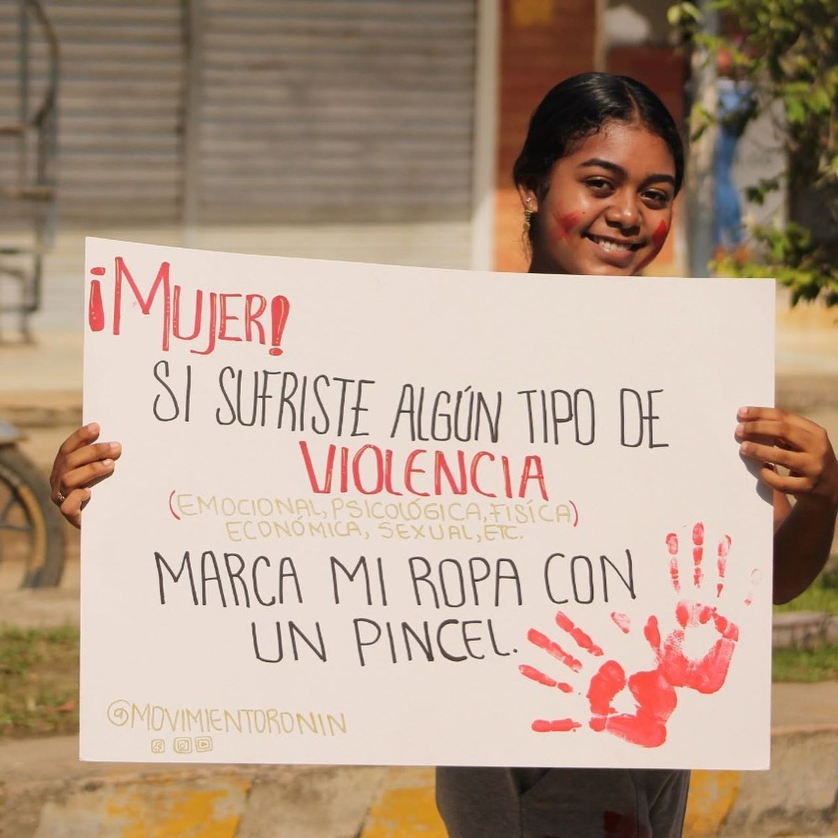 Fotografía de una joven sonriente que sostiene un cartel que dice «¡Mujer! Si sufriste algún tipo de violencia, marca mi ropa con un pincel», con la huella de una mano roja.